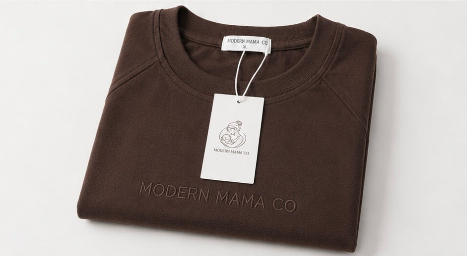 A folded dark brown crewneck sweatshirt with 'MODERN MAMA CO' debossed on the front, featuring a white hangtag and neck label.
